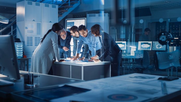Engineers Meeting in Technology Research Laboratory: Engineers, Scientists and Developers Gathered Around Illuminated Conference Table, Talking and Finding Solution, Inspecting and Analysing Industrial Engine Design.